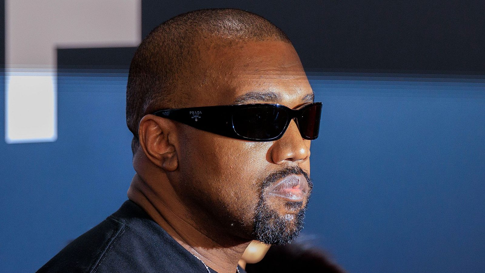 Primer plano de un hombre de raza negra con gafas de sol oscuras y camiseta negra. Tiene la cabeza rapada y una barba incipiente.