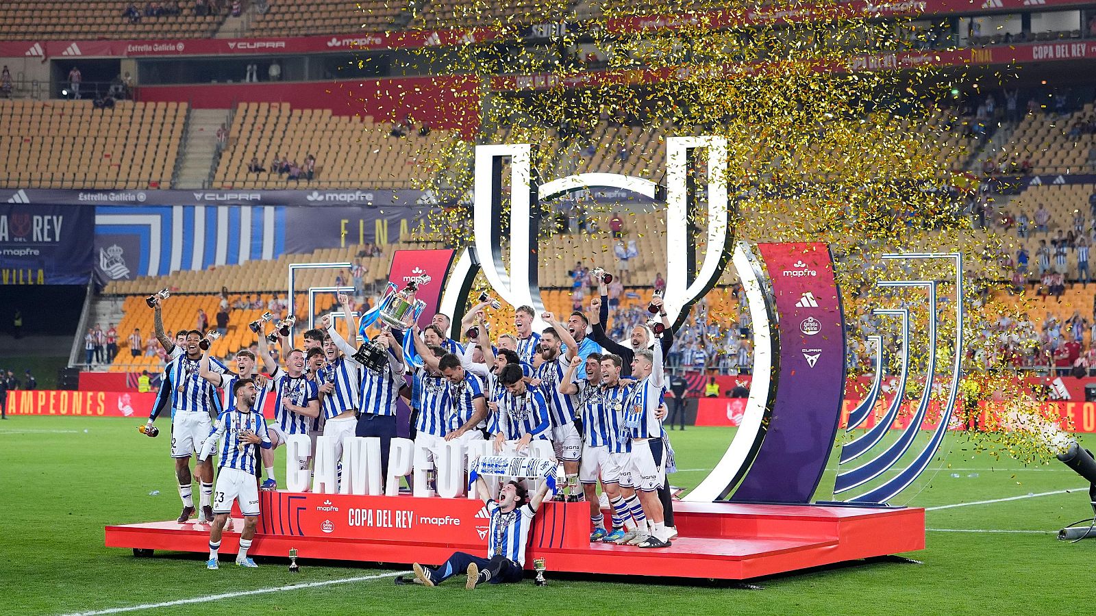 El equipo de la Real Sociedad celebra la victoria de la Copa del Rey en el campo, con el trofeo en alto y confeti cayendo, rodeados de aficionados en las gradas.