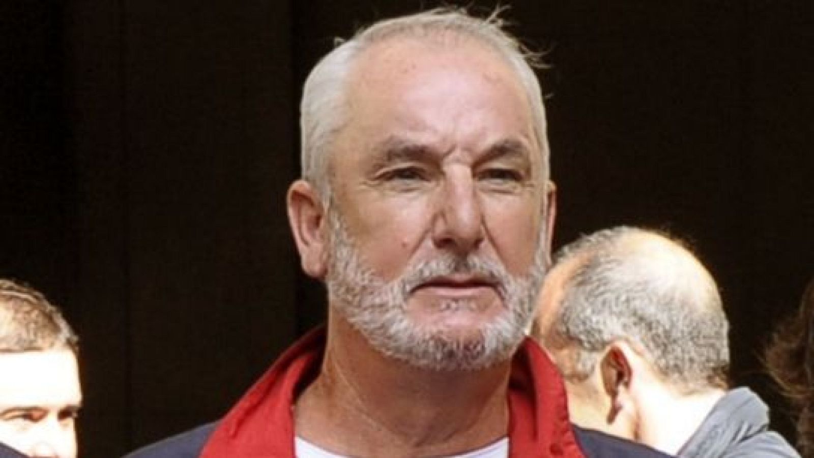 La fotografía presenta a un hombre de mediana edad con cabello y barba canosos, mostrando arrugas y una expresión seria. Su vestimenta incluye una chaqueta oscura con un cuello rojo.