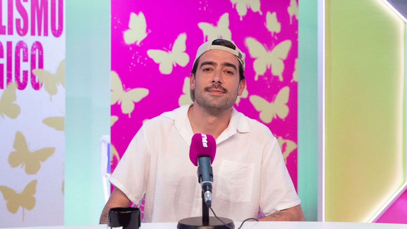 Marc Biarnes, presentador de radio, posa en un estudio con un micrófono magenta "PRPL" y un fondo de paneles rosas y mariposas.