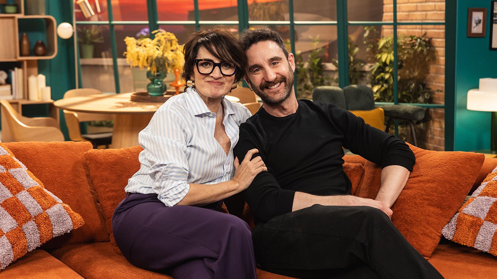 Dani Rovira i Sílvia Abril, somrients, comparteixen sofà. Ella llueix camisa de ratlles i ulleres, ell vesteix de negre, en una estança amb taula rodona i estanteria.