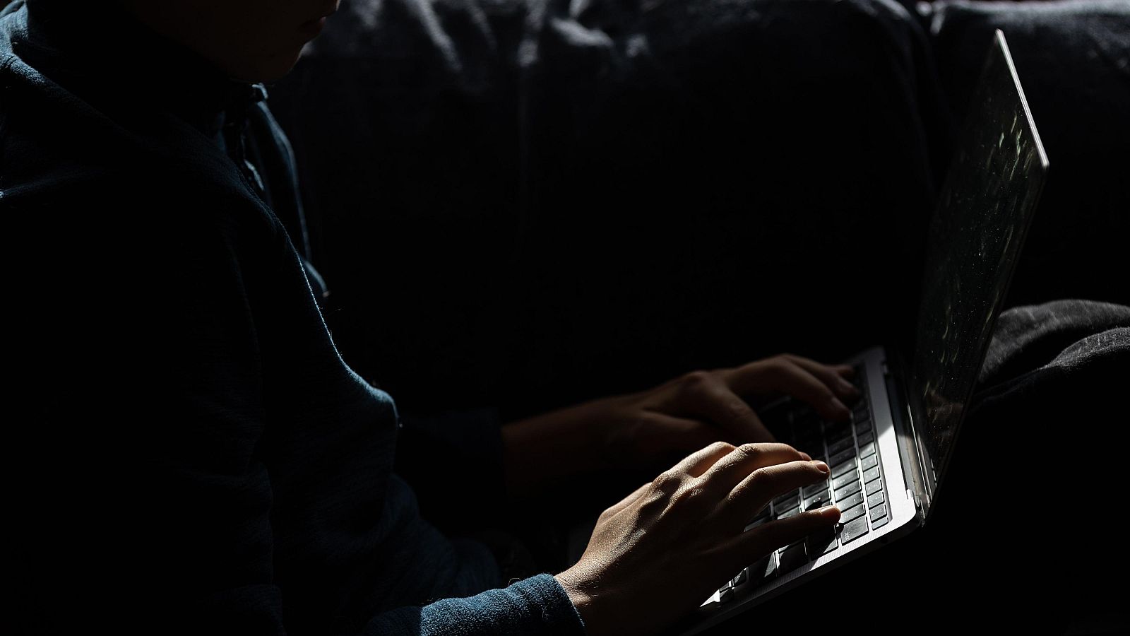 Fotografía de un chico con una sudadera azul mirando la pantalla de una computadora portátil en una habitación oscura