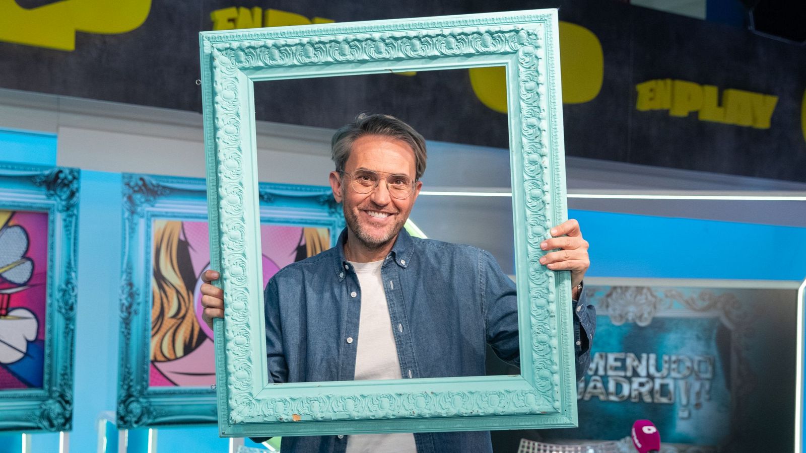 Máximo Huerta, con gafas y camisa vaquera, sonríe sosteniendo un marco turquesa en un plató de televisión. Se observan cuadros con ilustraciones y un rótulo con el título de un programa.