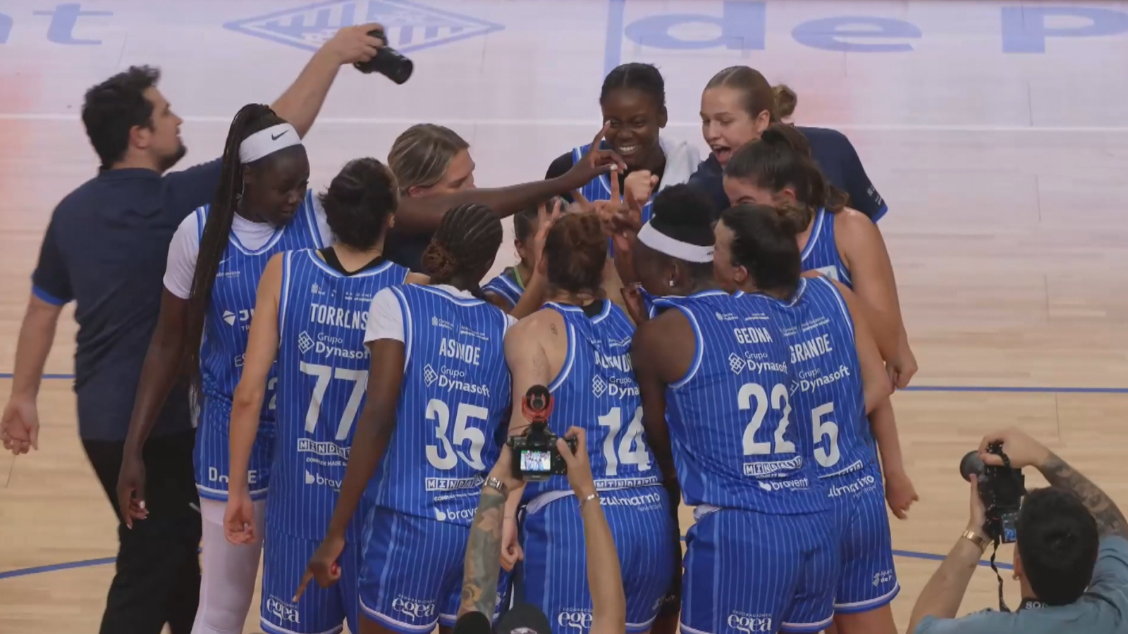 Un equipo de baloncesto femenino, con camisetas azules y rayas blancas, celebra una victoria en la cancha, abrazándose en un círculo. Se distinguen los números y nombres de algunas jugadoras en sus camisetas.