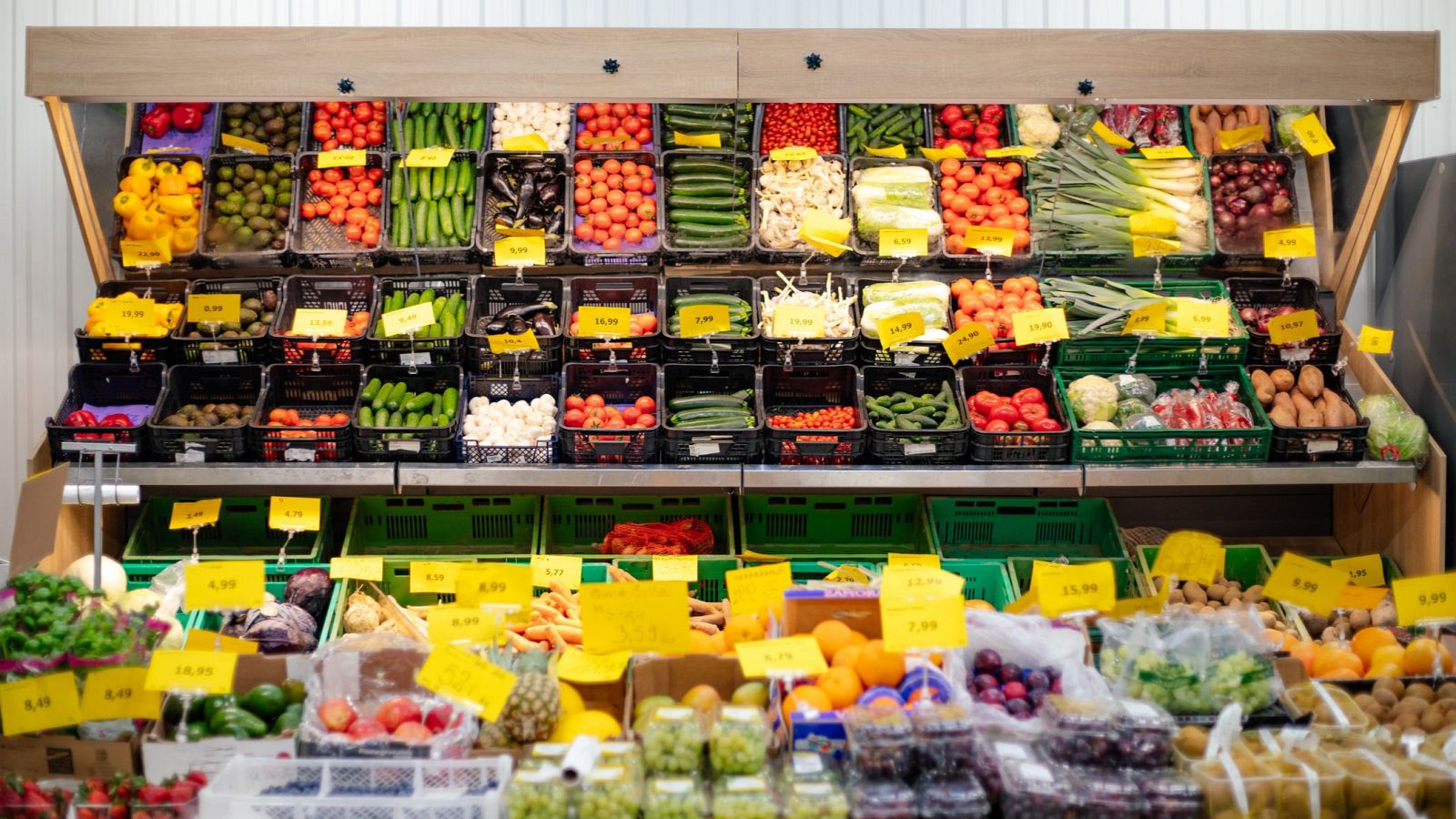 Expositor de madera con frutas y verduras frescas: pimientos, tomates, pepinos, berenjenas, champiñones, coliflor, lechugas, kiwis, patatas, limas, aguacates, naranjas, mandarinas, piñas. Etiquetas amarillas con precios.