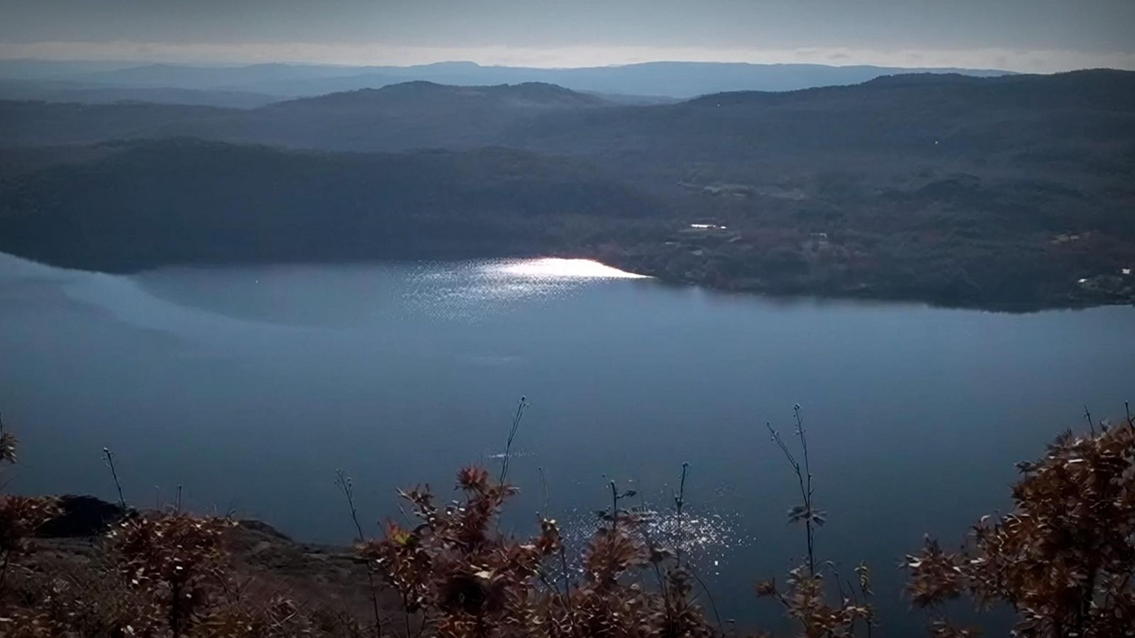 Paisaje gallego con ría, vegetación seca y montañas. Reflejo solar intenso en el agua. Vista elevada.