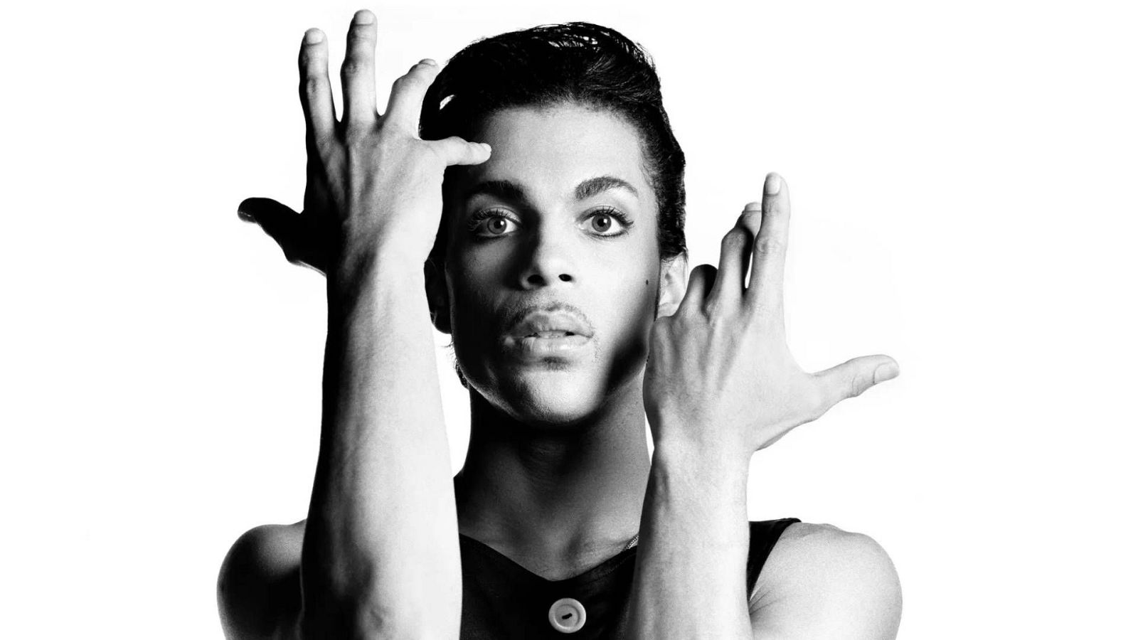 Un retrato en blanco y negro muestra a un hombre con el cabello peinado hacia atrás y una expresión intensa, con las manos cerca de su rostro y una camiseta sin mangas oscura.