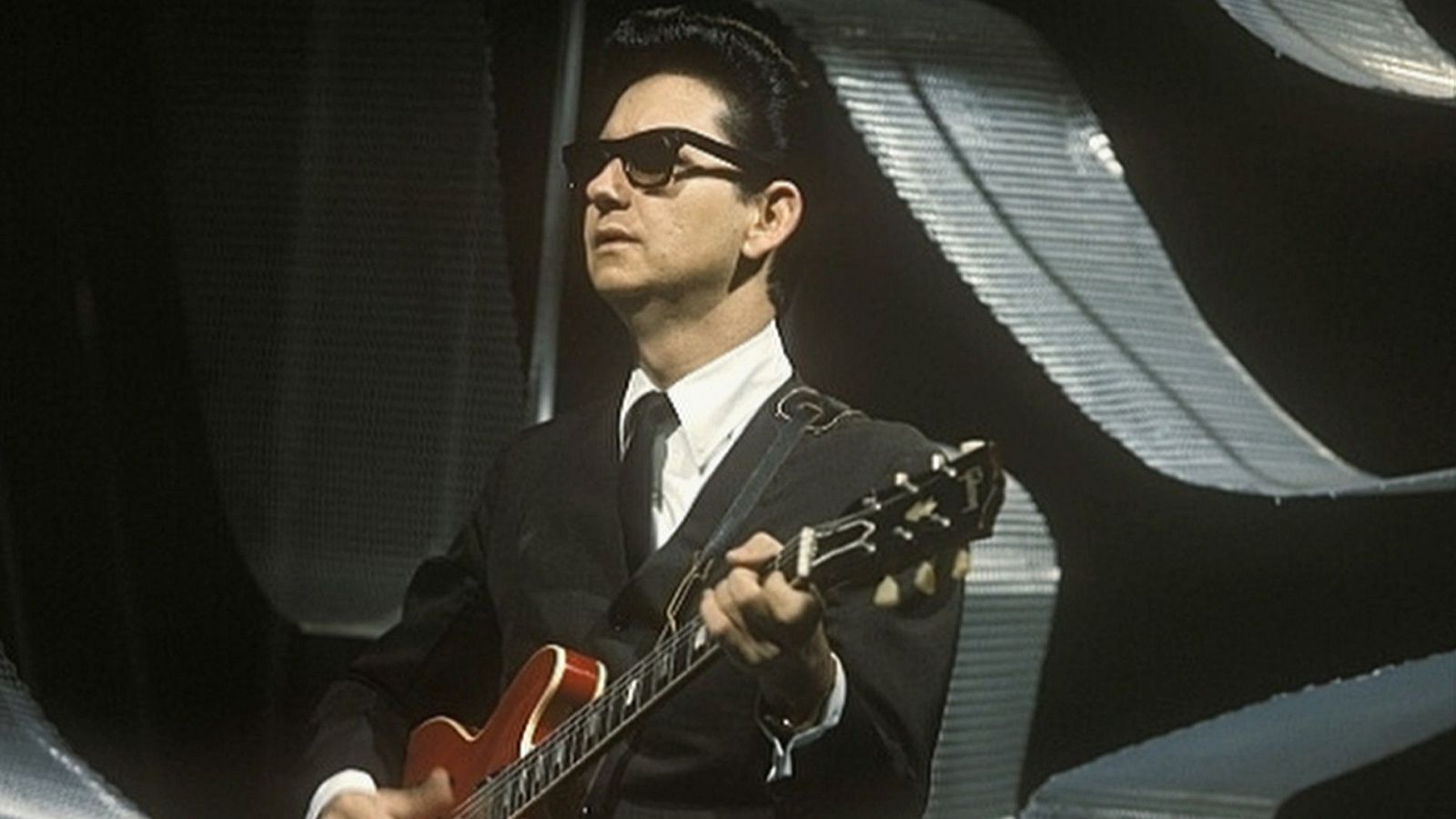 Roy Orbison, con gafas oscuras y traje, toca una guitarra eléctrica rojiza en un escenario con fondo texturizado y oscuro.