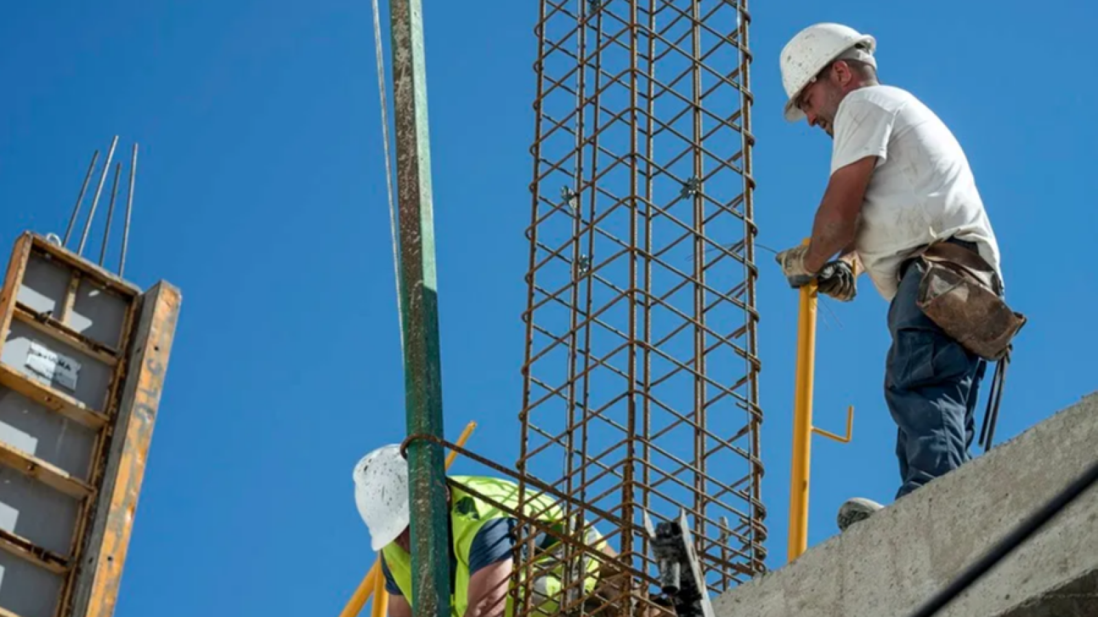 Un obrero con casco blanco y camisa blanca, atando alambres a una estructura de acero, mientras otro obrero con chaleco reflectante trabaja en la parte inferior, con el cielo azul de fondo.