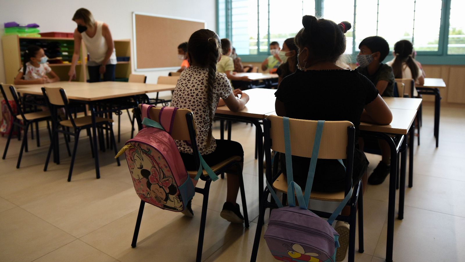 Grupo de niños y niñas de primaria sentados en sus pupitres atendiendo a una explicación en un aula escolar luminosa.
