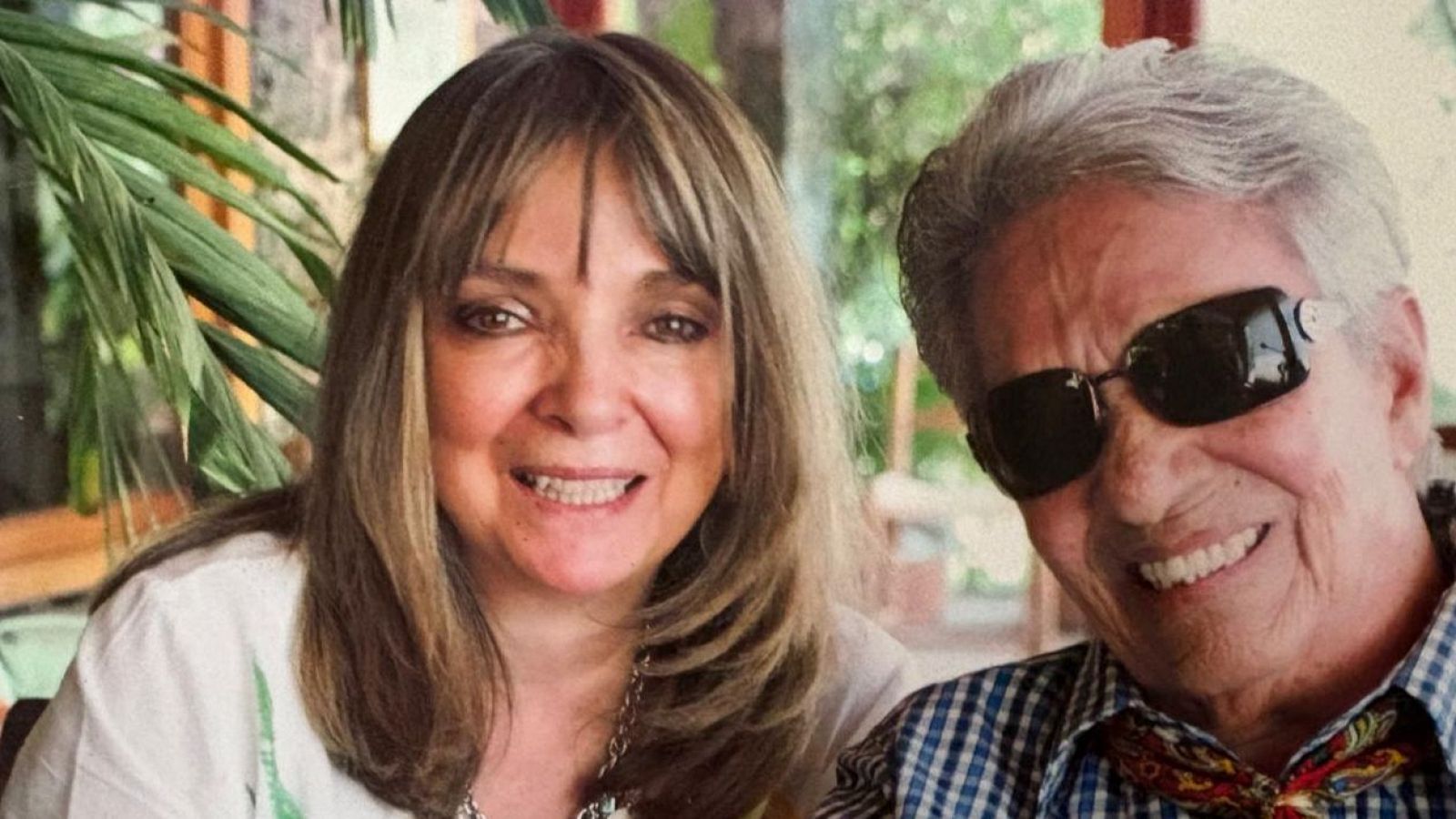 Arturo Martín, presentador de "Memoria de delfín", sonríe junto a una invitada. Ella lleva blusa blanca y collar plateado; él, camisa de cuadros y gafas de sol.