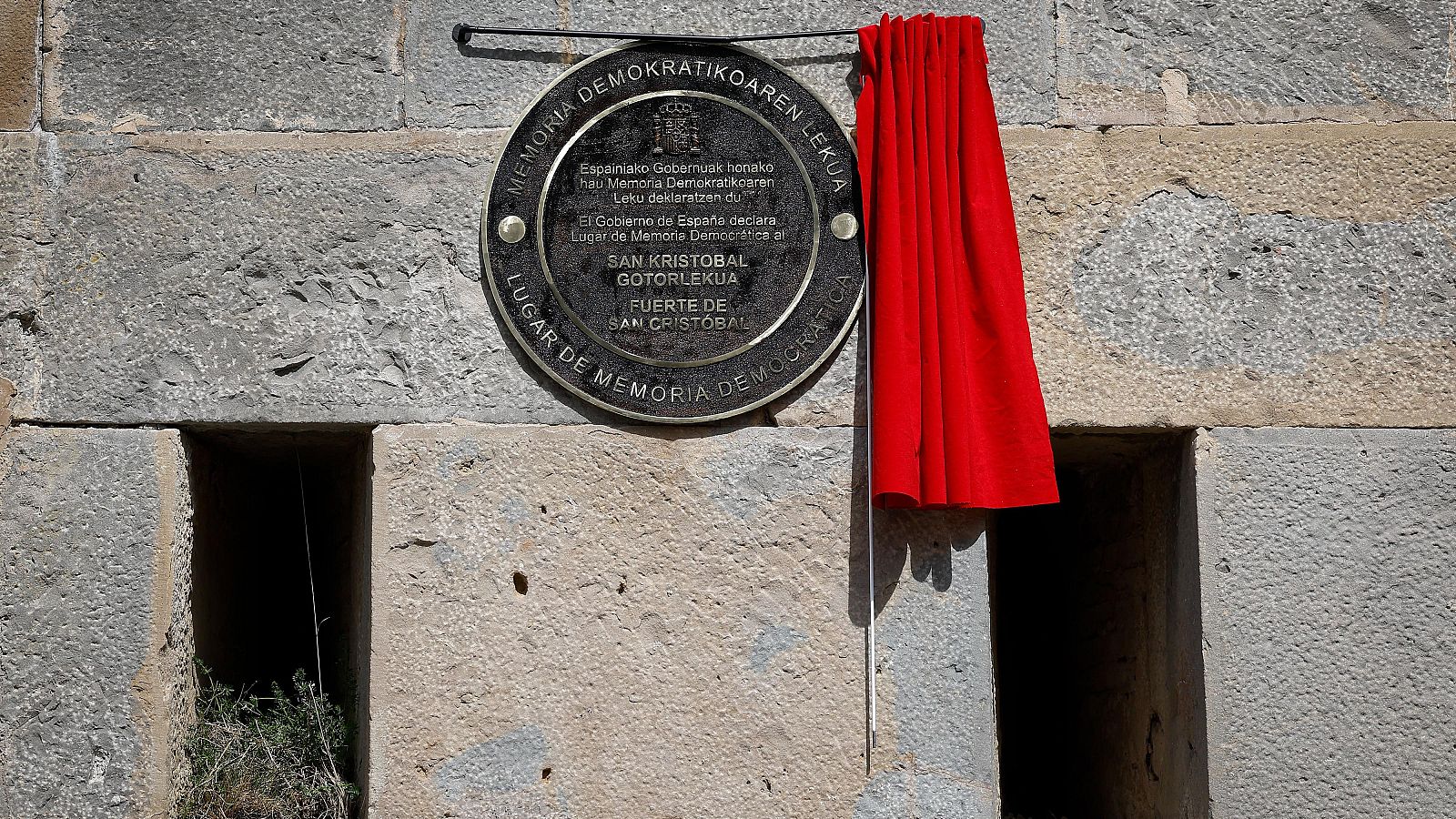 Placa bilingüe "LUGAR DE MEMORIA DEMOCRÁTICA" y "MEMORIA DEMOKRATIKOAREN LEKUA" con el Escudo de España en el Fuerte de San Cristóbal. Tela roja cubre parte de la placa.