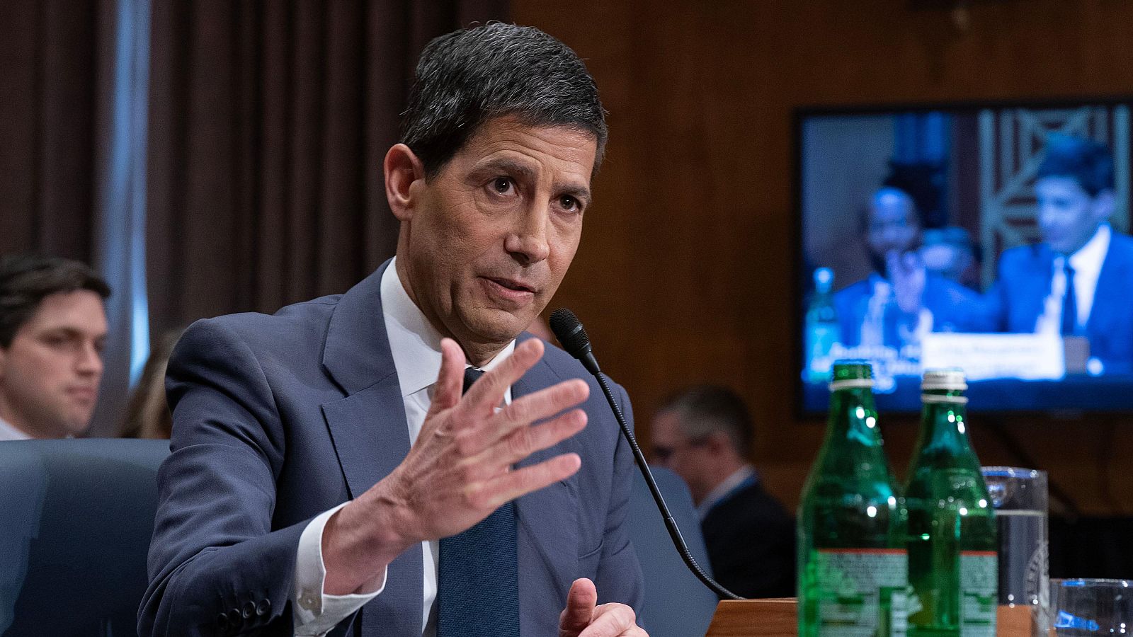 Kevin Warsh, candidato a la Fed, testifica con seriedad y gestos enfáticos. Viste traje azul oscuro y corbata. A su lado, botellas y un vaso. Pantalla al fondo muestra el evento.