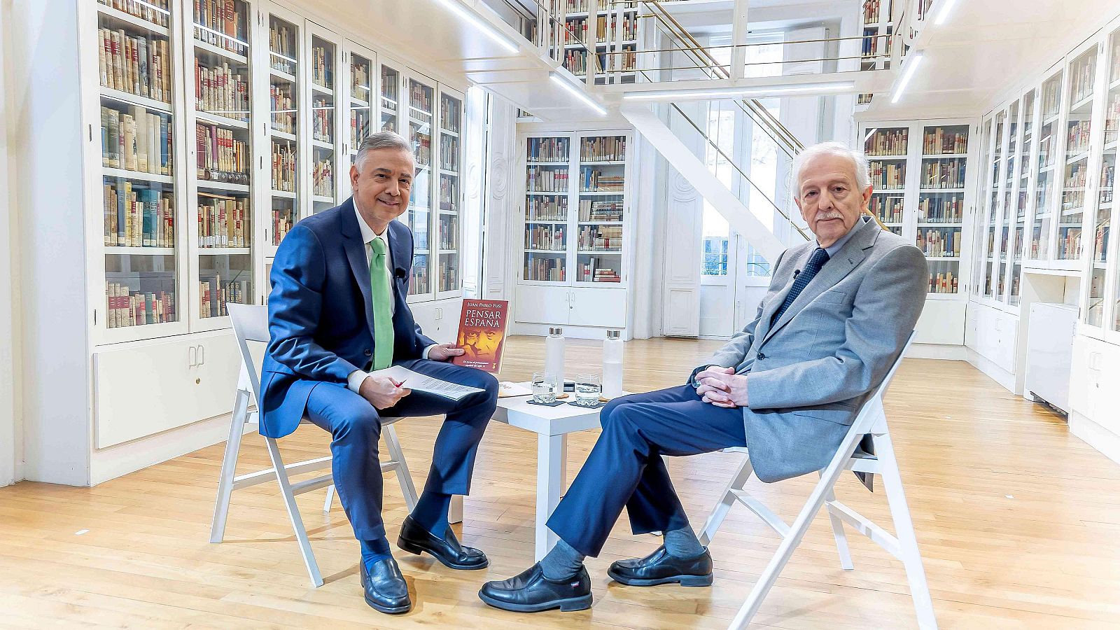 Jenaro Castro y Juan Pablo Fusi en una biblioteca, rodeados de libros. Castro sostiene "PENSAR ESPAÑA", Fusi tiene las manos entrelazadas. Hay botellas de agua y posavasos sobre una mesa.
