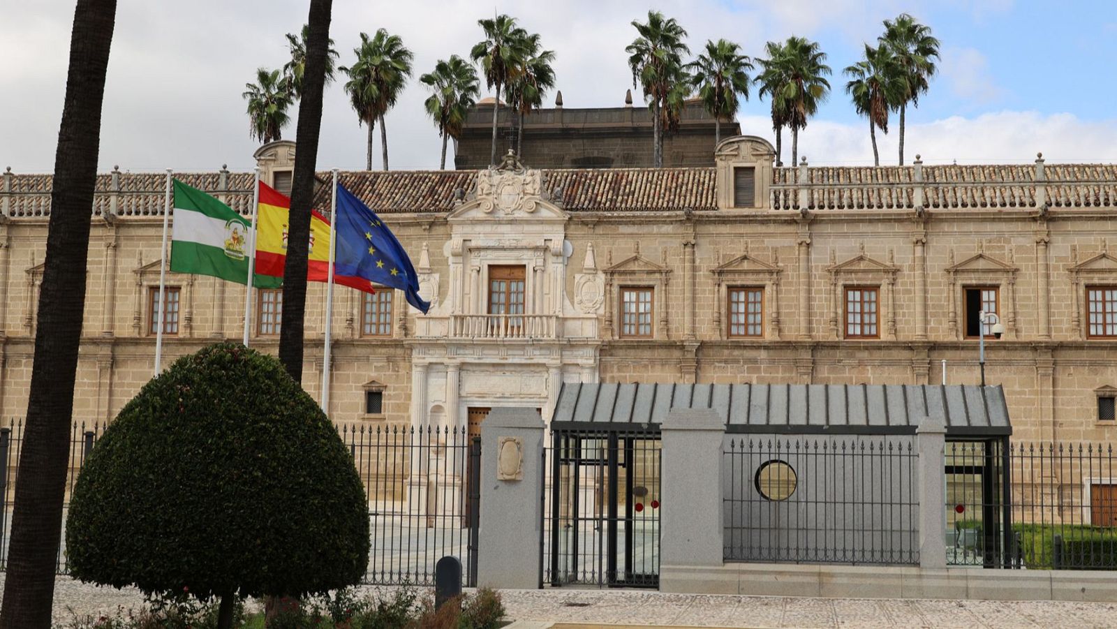 Imponente Palacio de San Telmo en Sevilla, sede de la Junta de Andalucía, con las banderas de Andalucía, España y la UE al frente, y palmeras elevándose sobre su tejado.