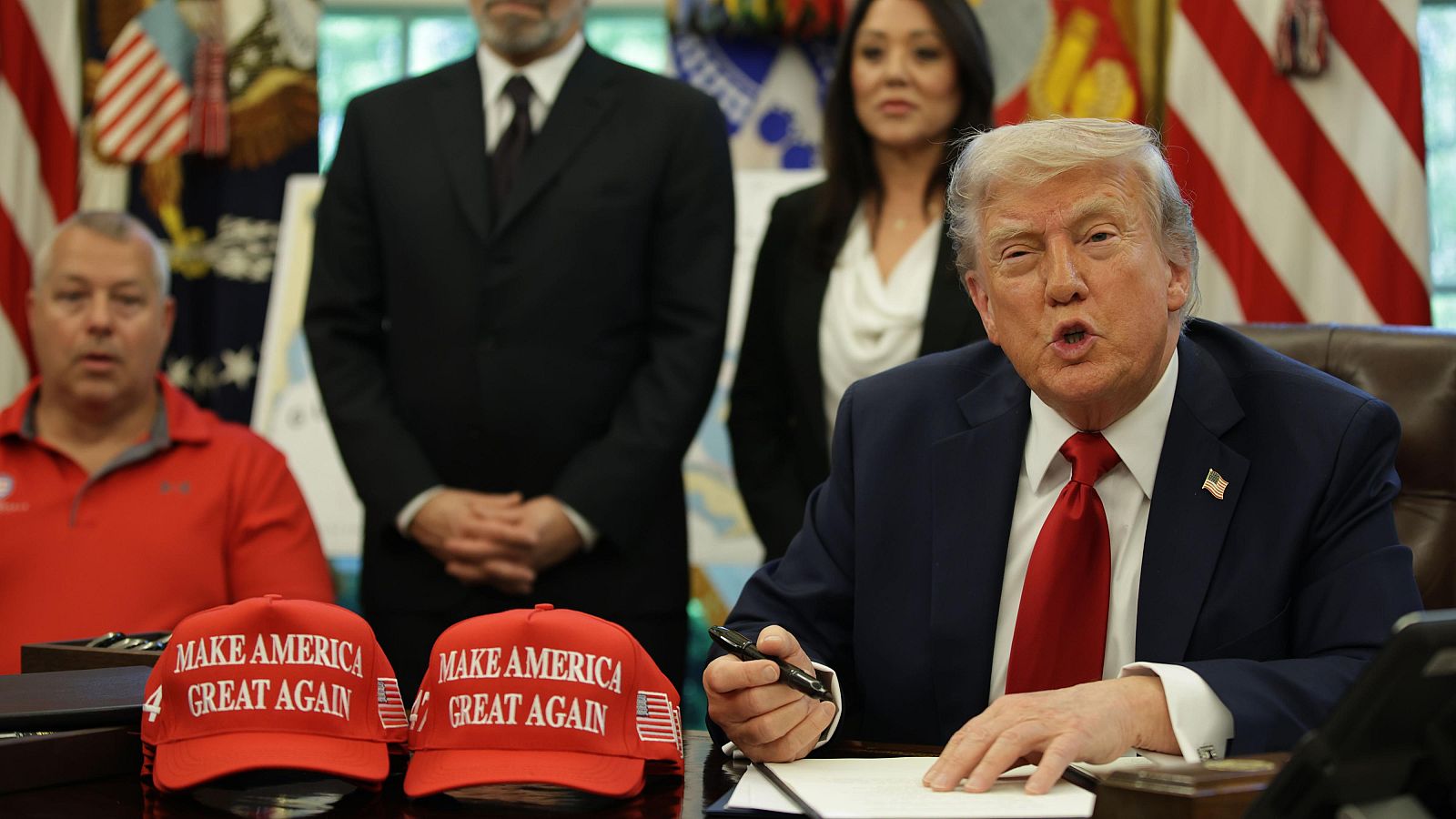 Donald Trump, con traje azul y corbata roja, sentado en un escritorio con gorras "MAKE AMERICA GREAT AGAIN" y banderas al fondo.