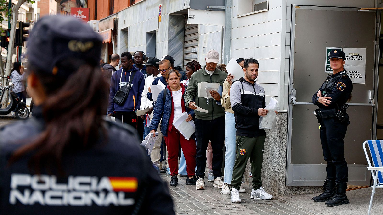 Agentes de la Policía Nacional supervisan una fila de personas con documentos frente a un edificio público. Se lee "POLICIA NACIONAL" y "NO SALIR SOLO EN CASOS DE EMERGENCIA".