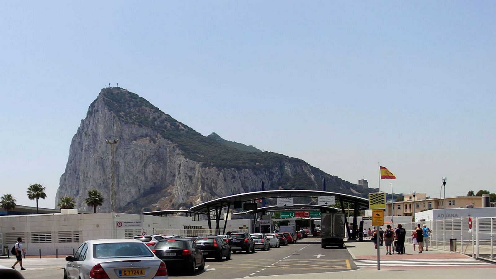 En la imagen se aprecia la frontera, con vehículos en espera y un paso de peatones. El Peñón de Gibraltar se alza imponente en el fondo, con una bandera española ondeando cerca.