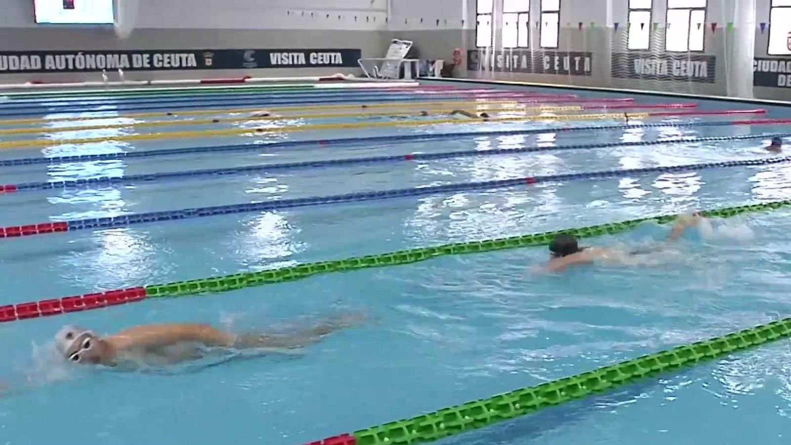 Piscina cubierta con nadadores a crol, corcheras de colores y textos promocionales de Ceuta en el fondo. Luz natural entra por grandes ventanales.