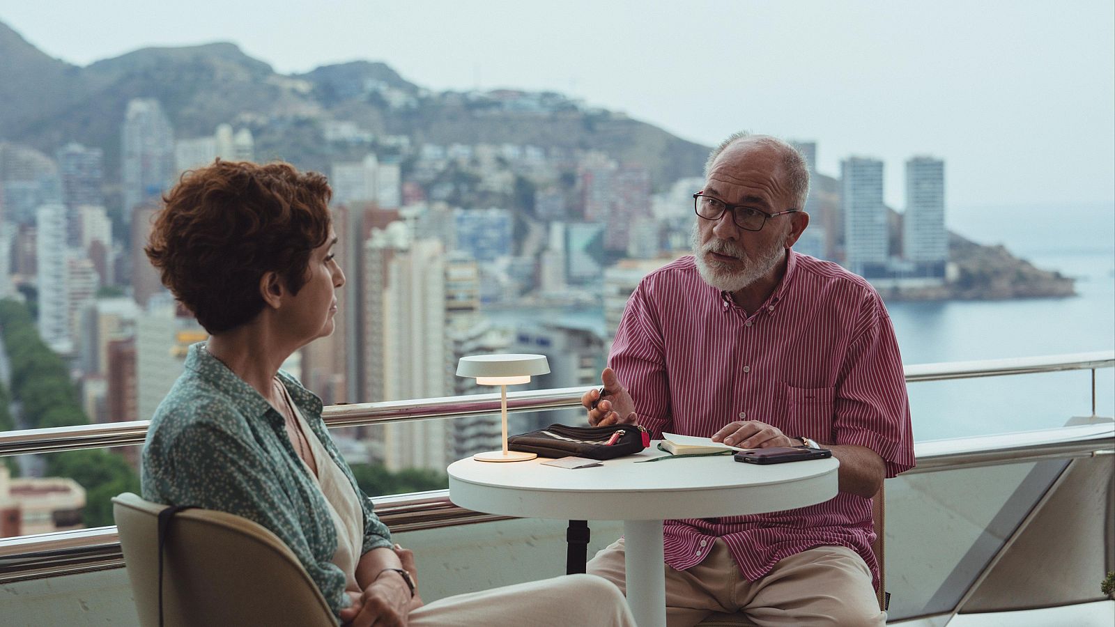 José Sacristán y María Galiana conversan en un balcón con vistas a una ciudad costera, rodeados de una lámpara, un cuaderno y un móvil.