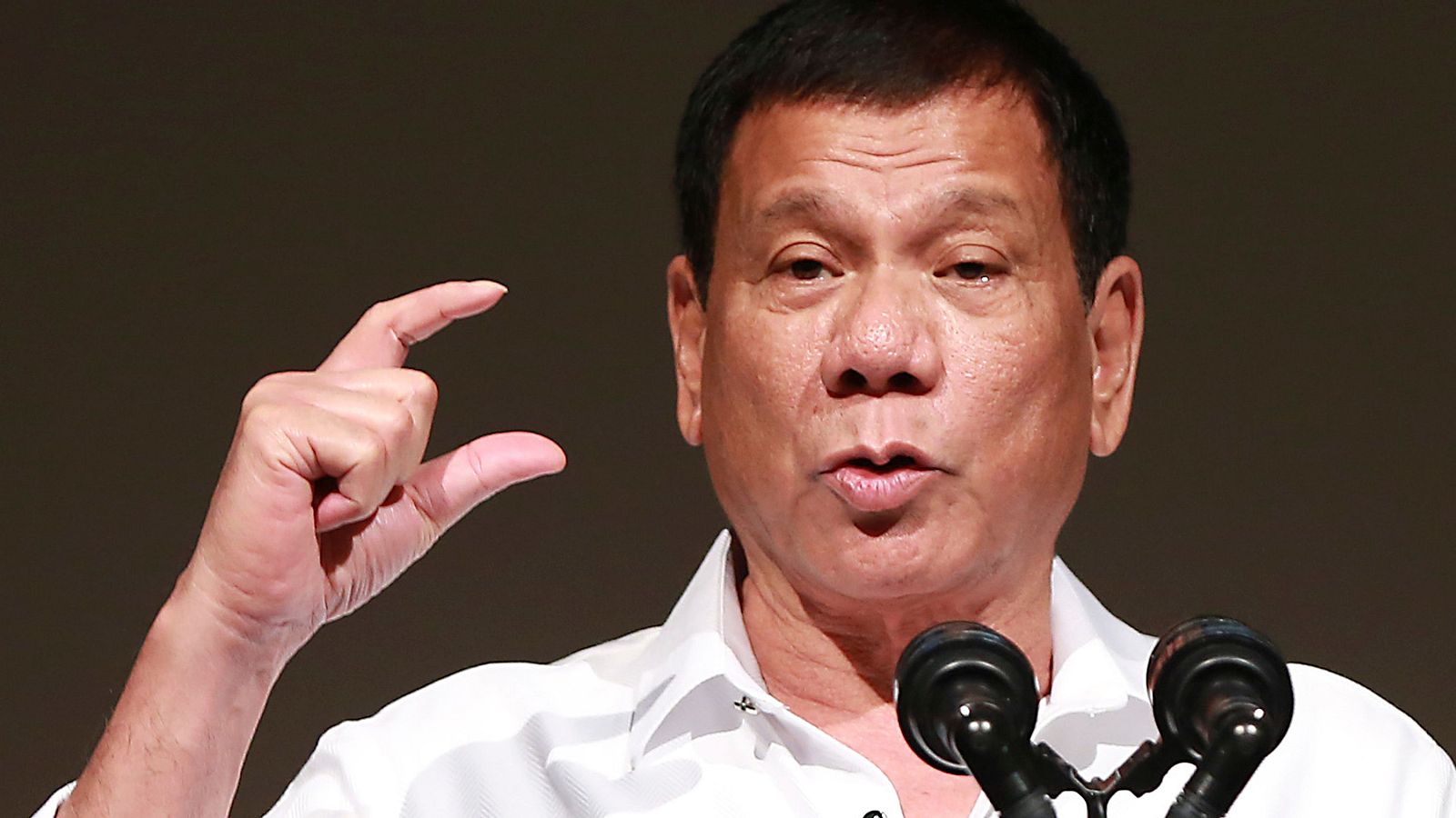 Primer plano de Rodrigo Duterte hablando, con la mano en gesto de énfasis y micrófonos al frente. Fondo oscuro y desenfocado.