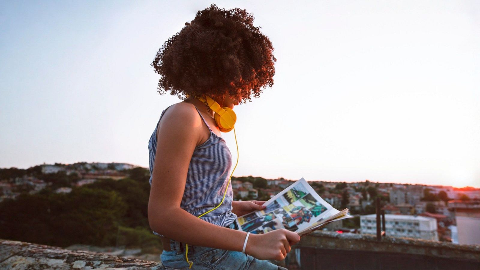 Joven con auriculares amarillos lee un cómic en un mirador al atardecer, con un paisaje urbano de fondo.