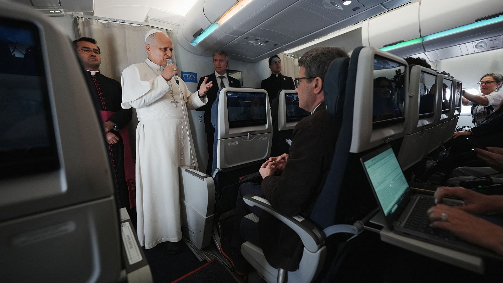 El Papa León XIV habla con periodistas en el pasillo de un avión, vestido con sotana blanca y solideo, sosteniendo un micrófono.