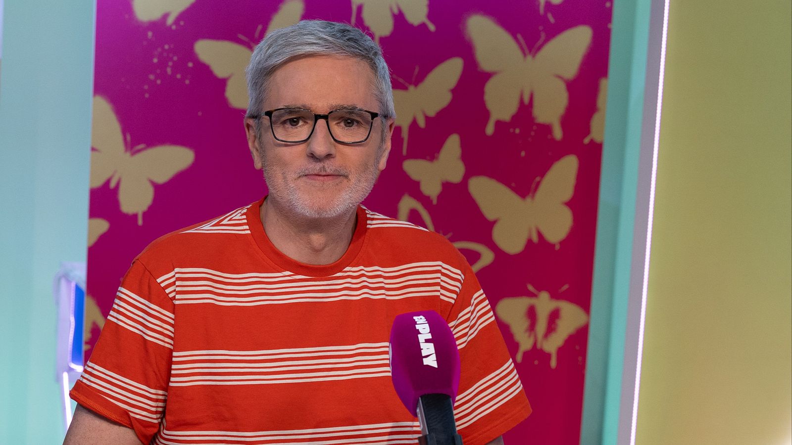 Mikel Iturriaga, con camiseta a rayas y gafas, frente a un micrófono morado 'SPLAY'. Fondo dividido en paredes fucsia con mariposas doradas y verde claro.