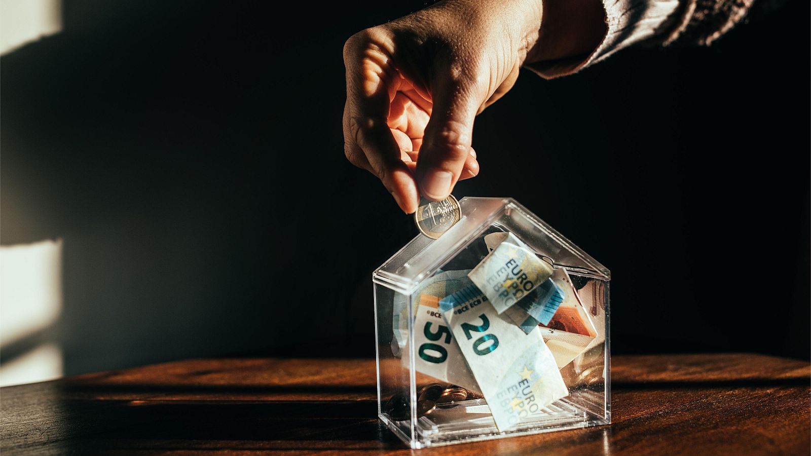Mano depositando moneda en hucha transparente con forma de casa, conteniendo billetes de euro y monedas. Escena de ahorro.