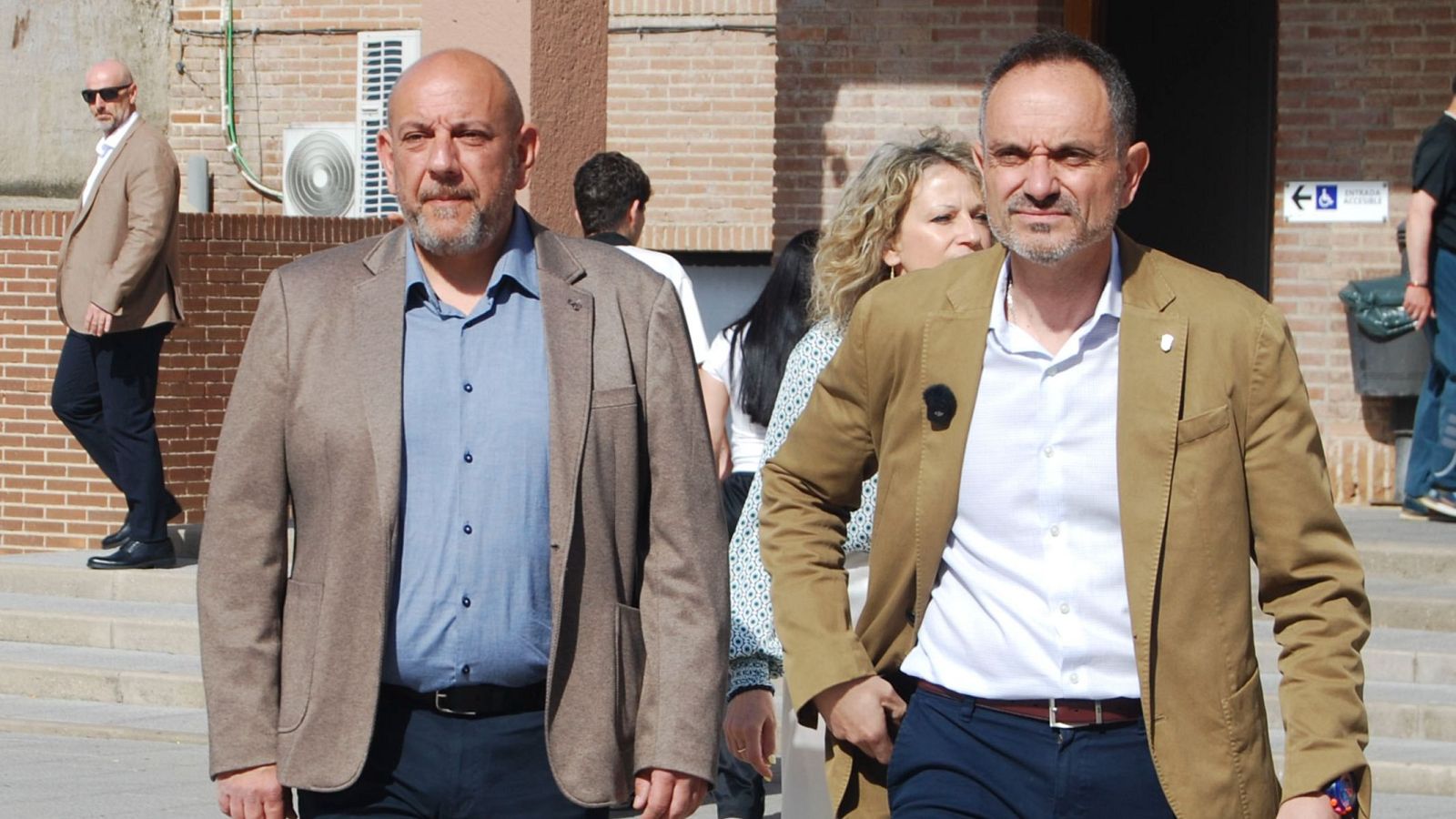 Manuel Bautista, alcalde de Móstoles, y Gabriel Monteserín, concejal de Seguridad, caminan en un día soleado frente a una pared de ladrillo con señalización.