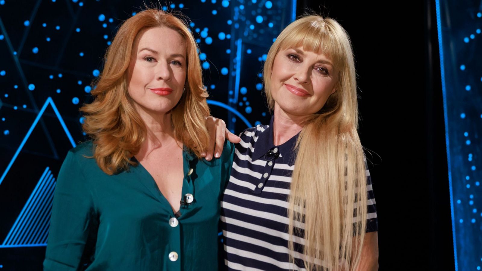 Lucía Quintana y Cayetana Guillén Cuervo posan sonrientes, una con vestido verde azulado y cabello pelirrojo, la otra con polo a rayas y cabello rubio, ante un fondo oscuro con luces azules.