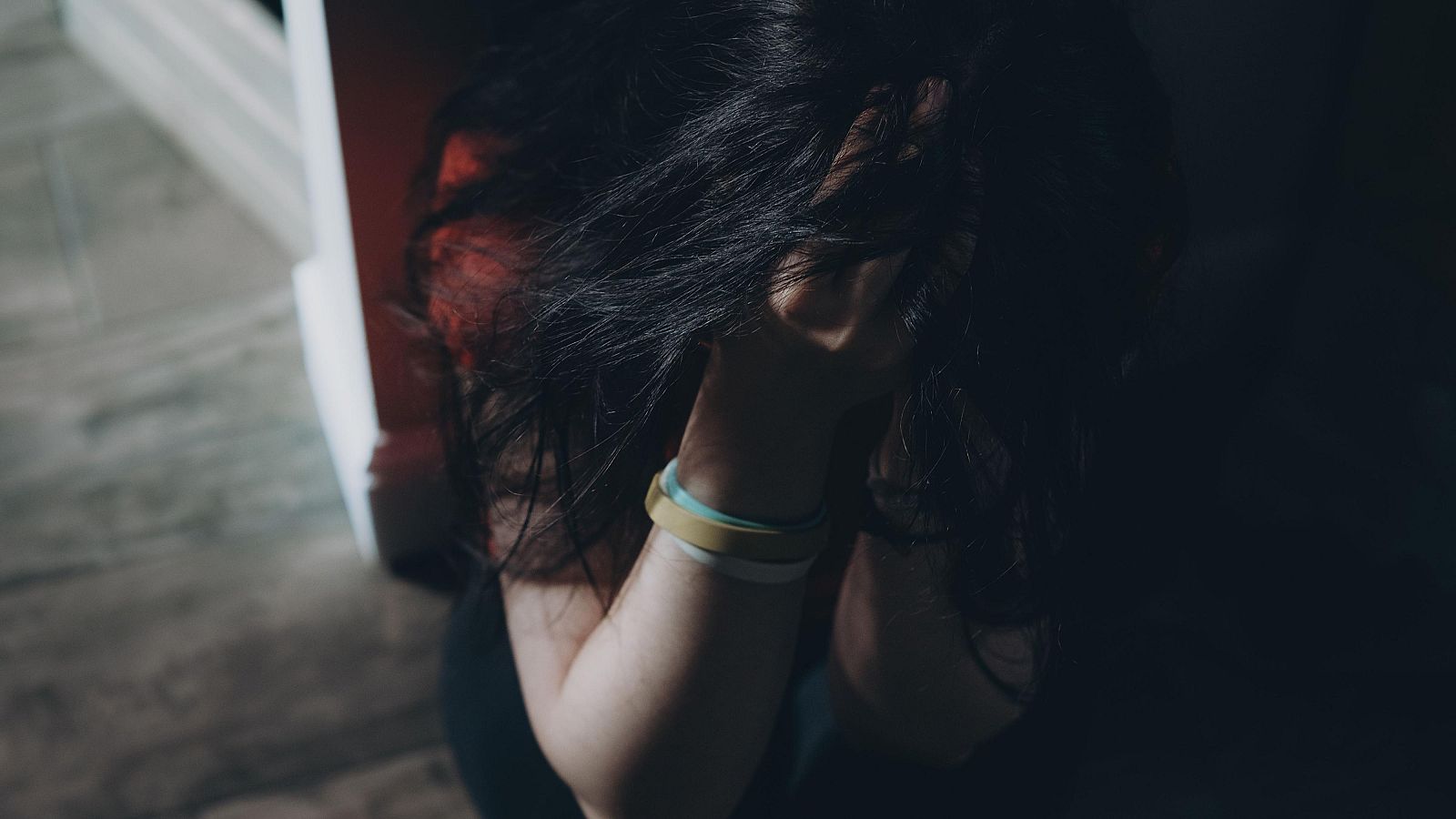 Fotografía de una adolescente angustiada se sienta en el suelo y se sujeta la cabeza, vestida con una camiseta roja y pulseras en la muñeca