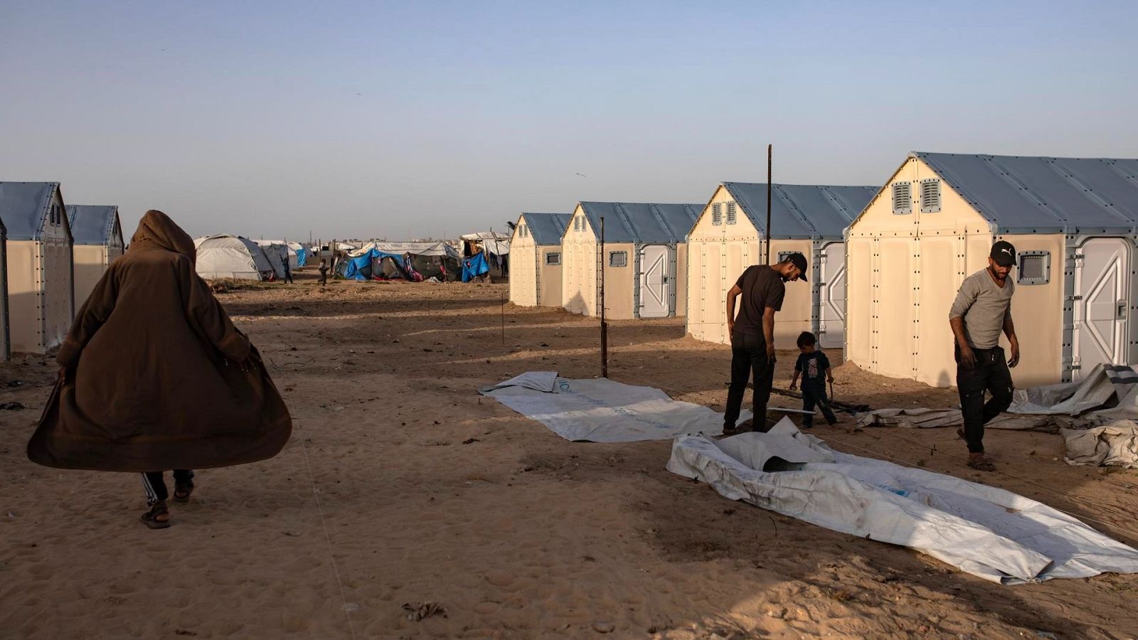 Personas caminan entre tiendas prefabricadas y tiendas de campaña en un campamento árido. Se aprecian lonas blancas en el suelo.