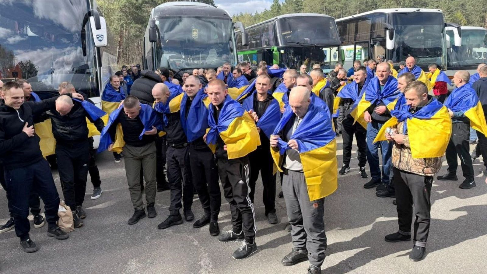 Hombres ucranianos liberados como prisioneros de guerra, envueltos en banderas de su país, se reúnen junto a autobuses. Expresiones de alivio y seriedad marcan la emotiva escena.