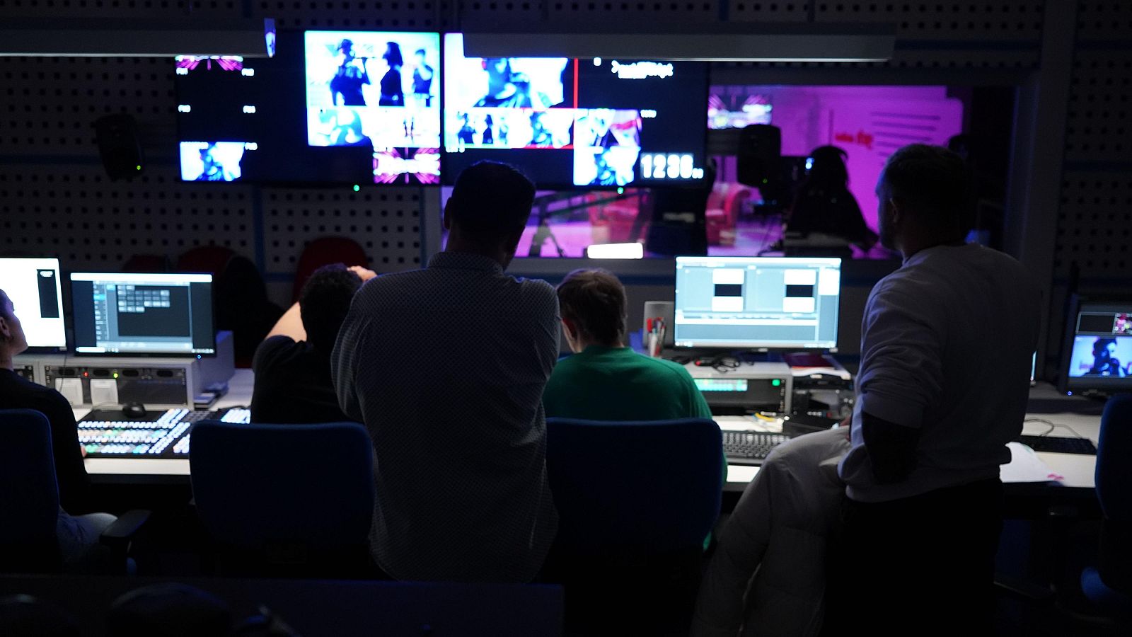 Sala de control con personas trabajando frente a monitores y pantallas de pared que muestran contenido multimedia. Se observa la hora "12:38." en una pantalla.