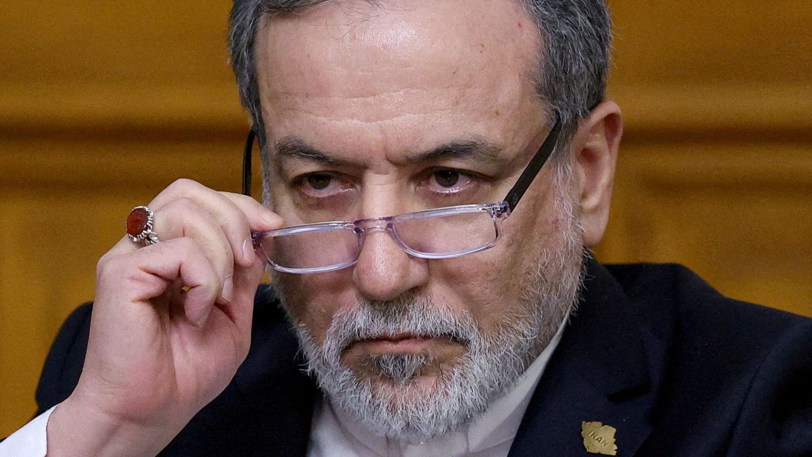 Abbas Araqchi, ministro de Asuntos Exteriores iraní, en rueda de prensa. Sostiene gafas, lleva anillo y pin "IRAN". Fondo ocre desenfocado.