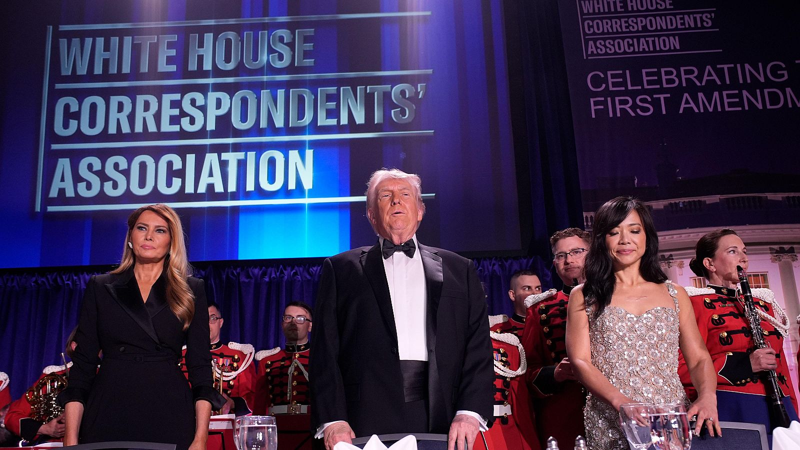 Donald Trump, Melania Trump y una mujer asiática en la cena de la Asociación de Corresponsales de la Casa Blanca. Pantallas con "WHITE HOUSE CORRESPONDENTS' ASSOCIATION" y "CELEBRATING THE FIRST AMENDMENT".