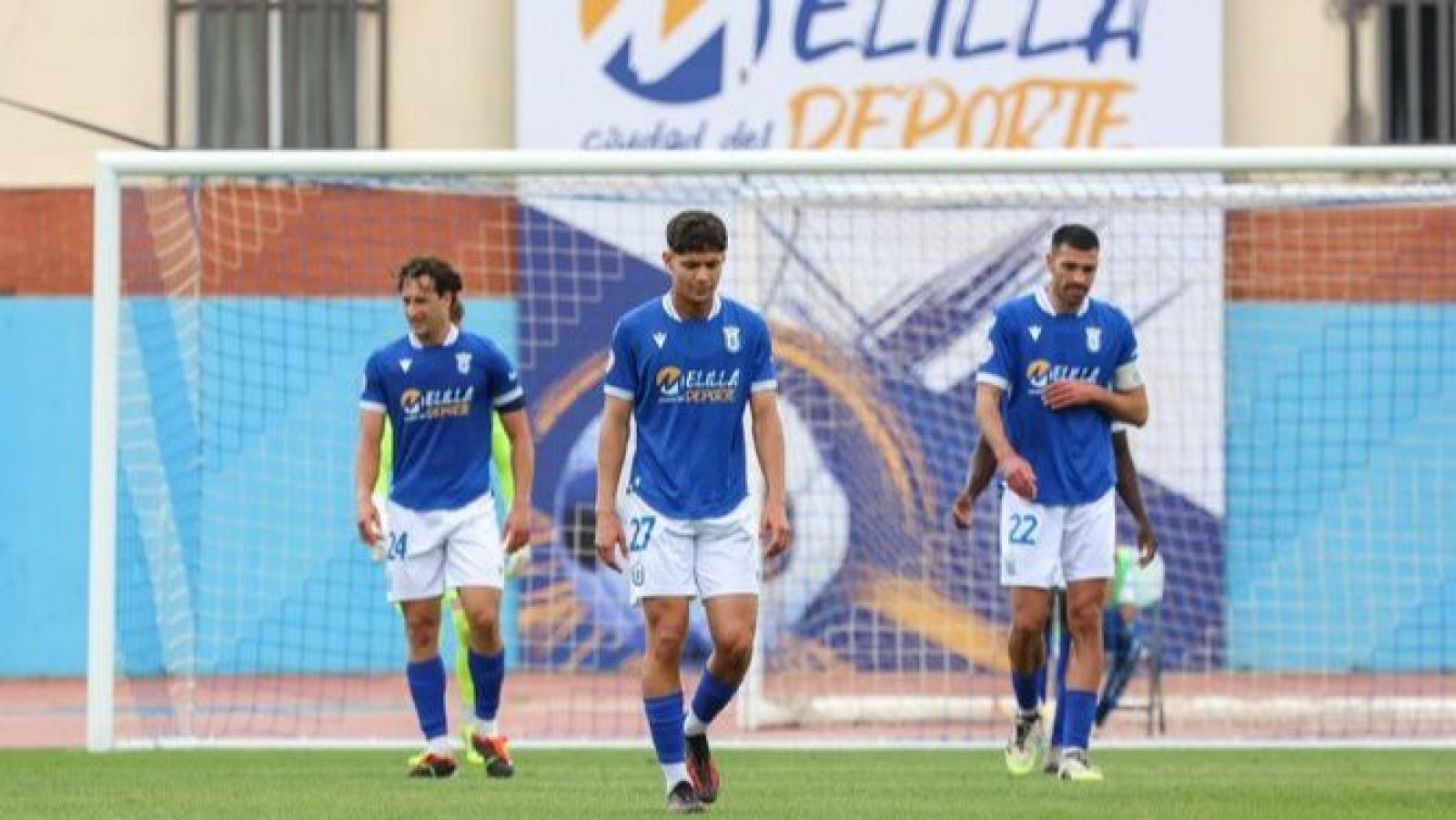 Jugadores de la UD Melilla con dorsales 24, 27 y 22, equipados con "MELILLA DEPORTE", en un campo de césped con portería y mural "MELILLA Ciudad del DEPORTE".