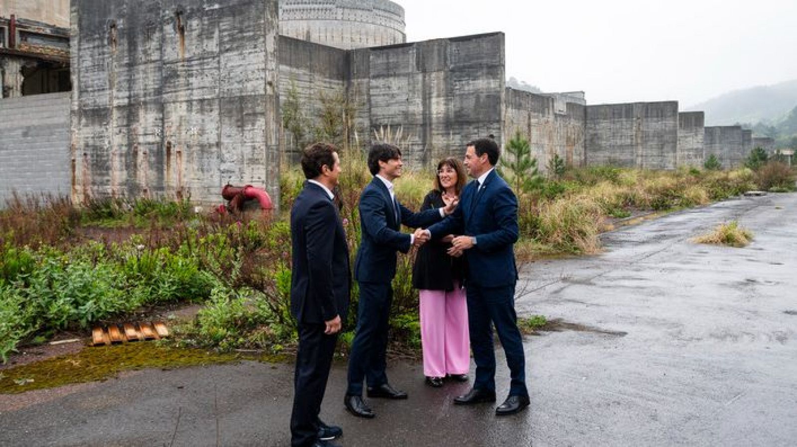 Imanol Pradales, Lehendakari, saluda a un hombre en la central nuclear de Lemoiz. Una mujer y otro hombre observan el encuentro. El paisaje industrial de la central nuclear se alza al fondo.