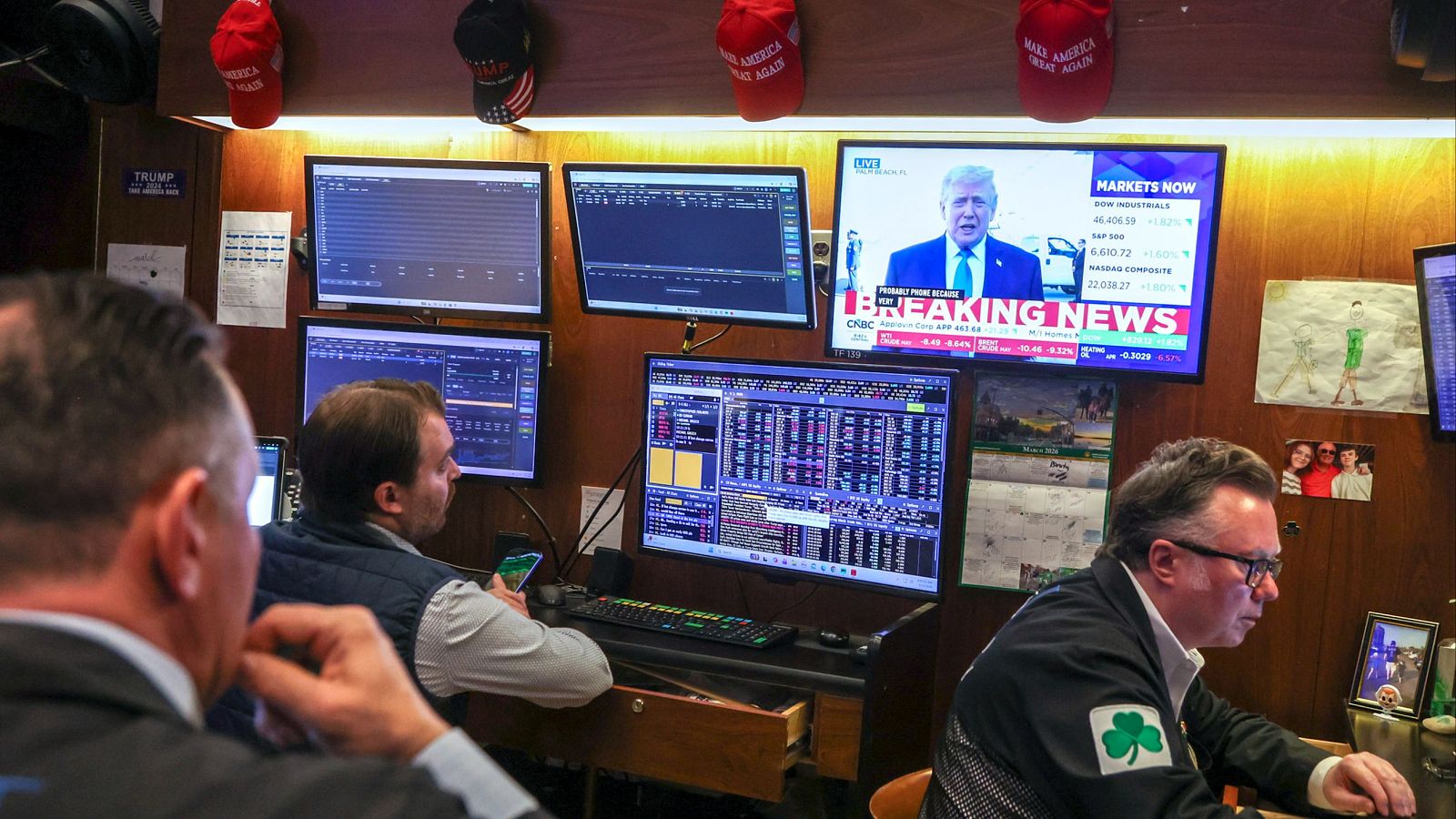 Tres hombres observan múltiples pantallas con información financiera y noticias. En una de ellas, se muestra a Donald Trump en un noticiero con el titular "BREAKING NEWS".