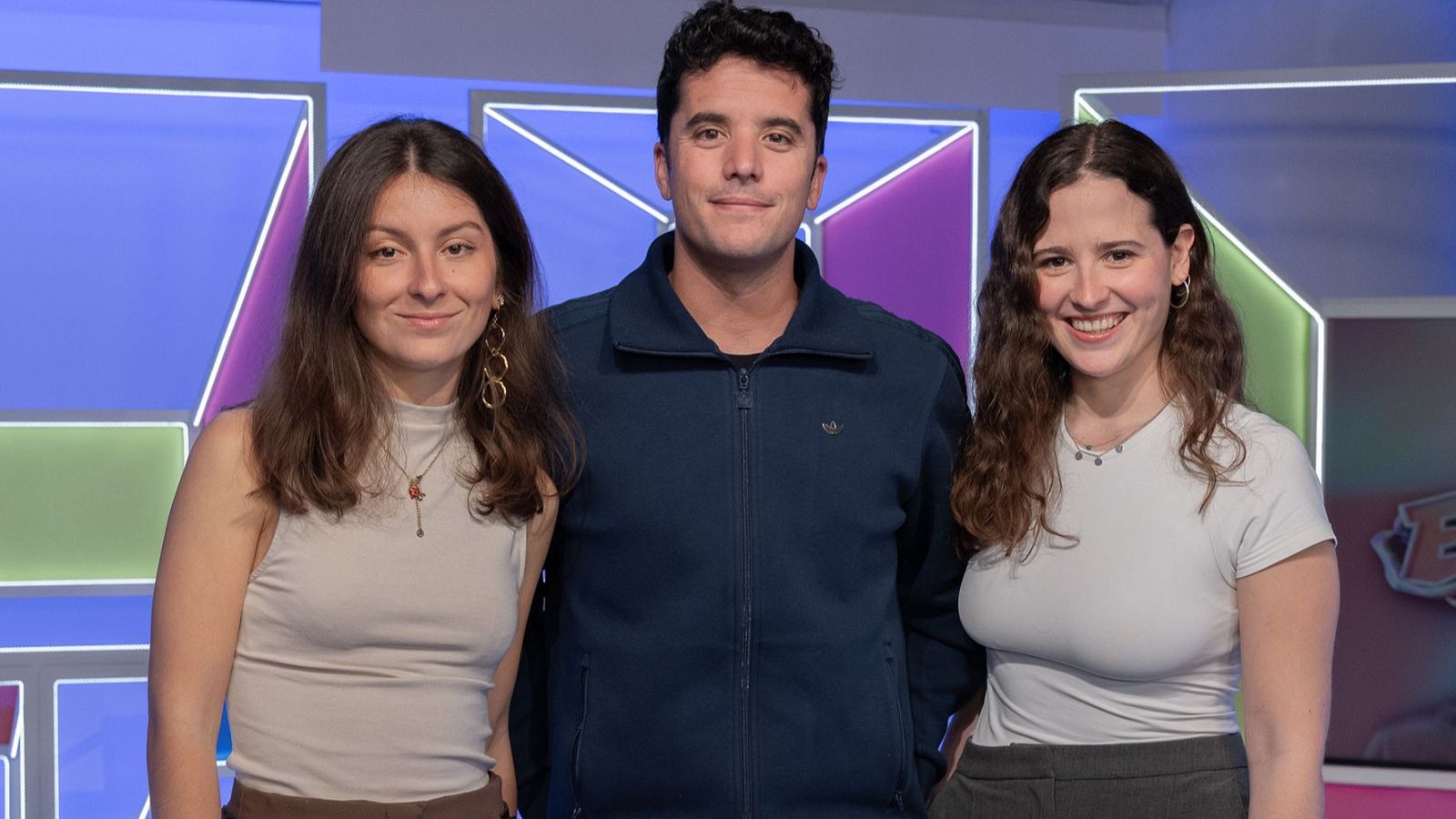 Nanísimo, Mar Manrique y Marina Enrich posan sonrientes en un estudio con fondo de neón. Nanísimo lleva chaqueta azul marino, Mar Manrique camiseta beige y Marina Enrich camiseta blanca.