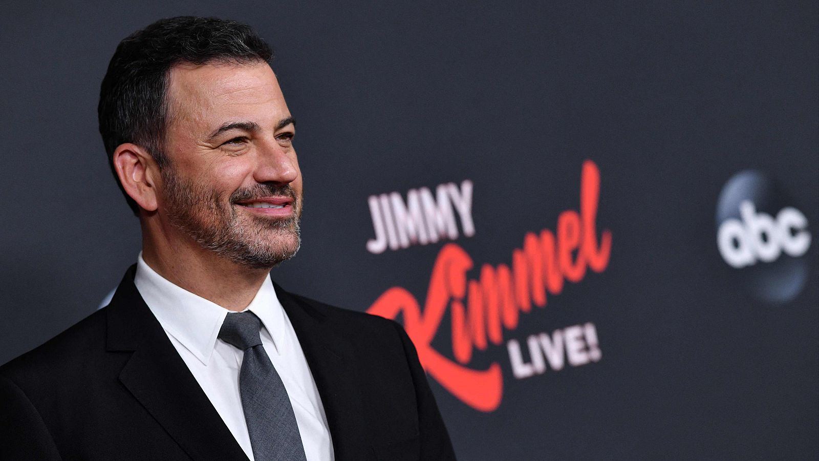 Jimmy Kimmel sonríe a la cámara, vestido formalmente, con el nombre de su programa "JIMMY Kimmel LIVE!" y el logo de "abc" en el fondo.