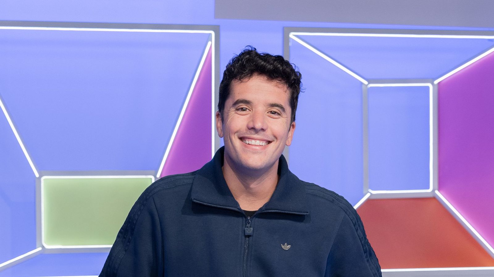 Nanísimo sonríe a la cámara con chaqueta azul y logo de Adidas, en un estudio de TV con paneles de colores y luces.