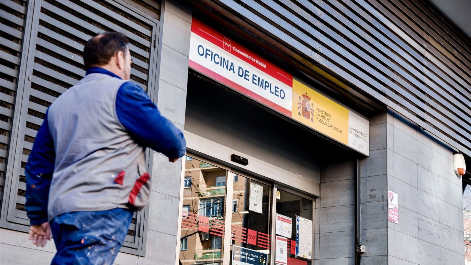 Persona con ropa de trabajo entrando en una oficina de empleo moderna con fachada de paneles metálicos y puerta de cristal. Cartel con nombre de la oficina y logotipos visibles.