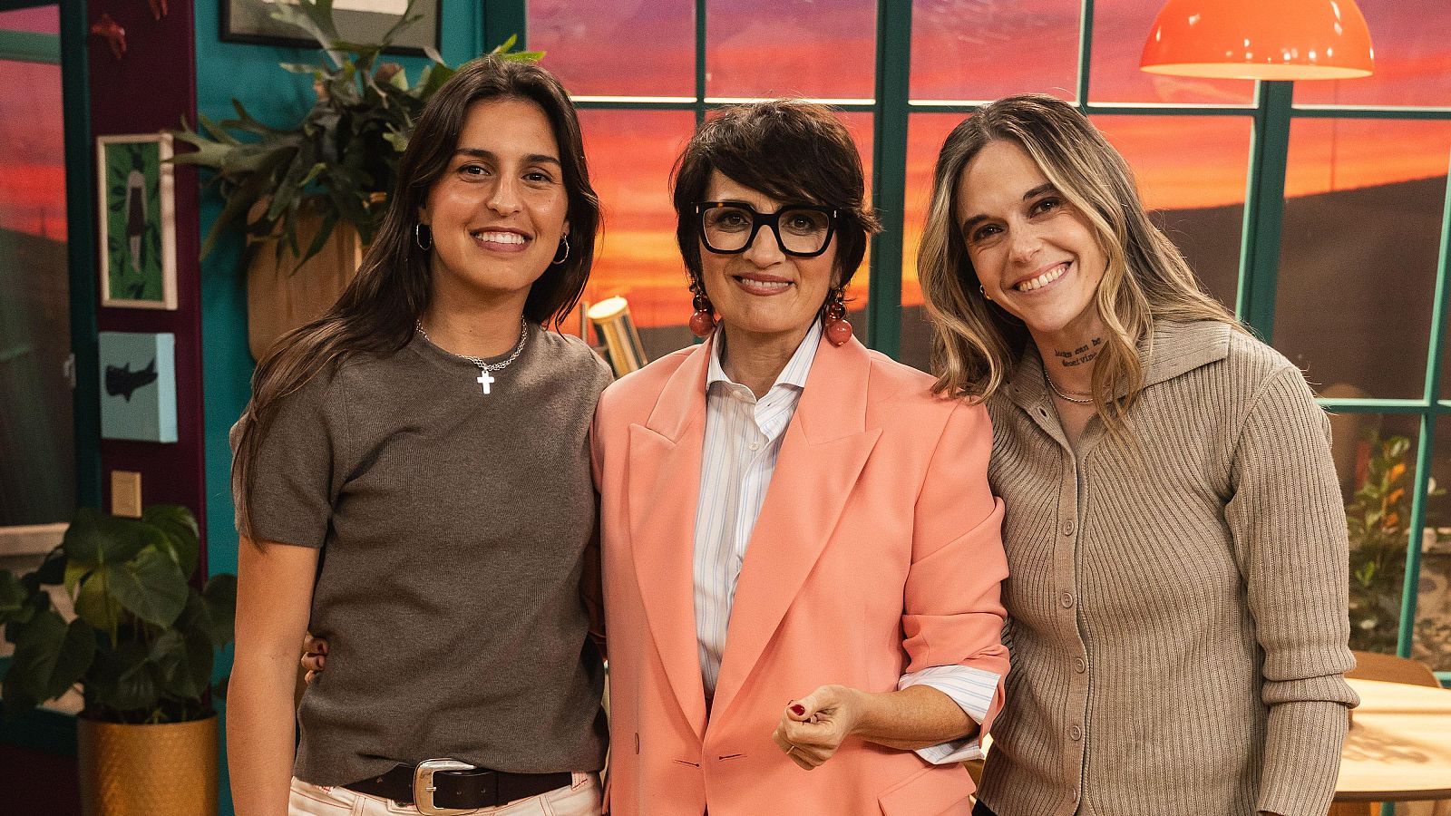 Sílvia Abril, Mapi León y Kika Nazareth sonríen juntas en un estudio de televisión con un fondo de atardecer. Sílvia Abril viste americana salmón, Mapi León camiseta gris y Kika Nazareth cárdigan beige.