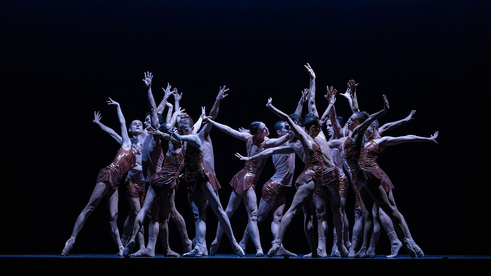Bailarines en trajes cobrizos ejecutan una coreografía expresiva sobre un escenario oscuro, bajo una iluminación dramática.