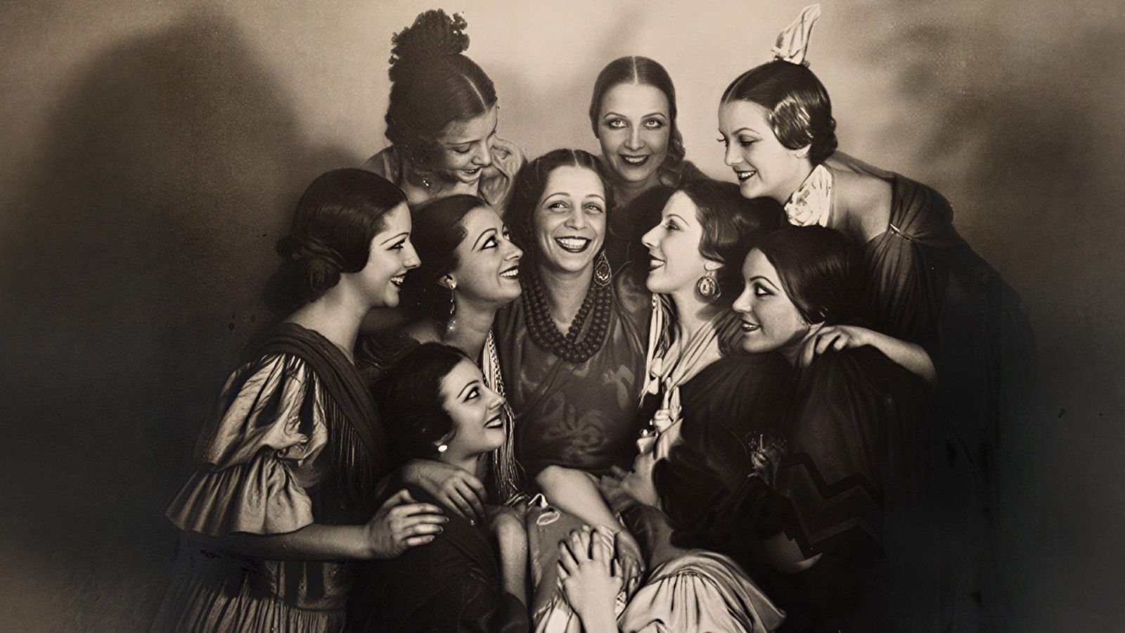 Antonia Mercé, "la Argentina", sonríe rodeada de ocho bailarinas con trajes de danza española. Fotografía histórica en blanco y negro de un grupo unido y feliz.