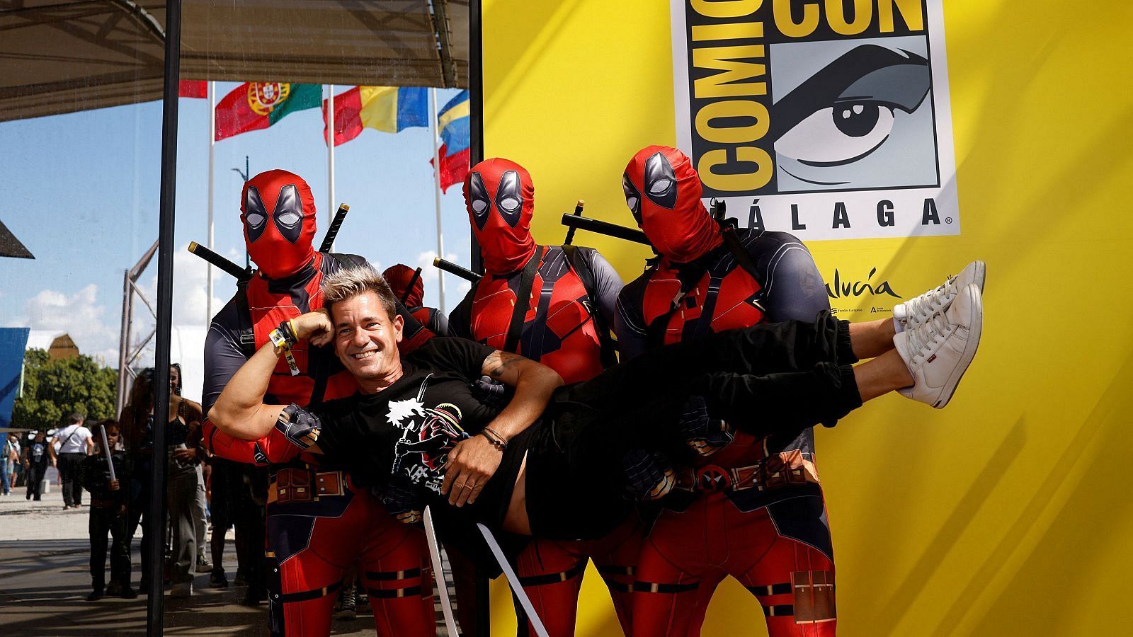Arturo Valls, sostenido por cosplayers de Deadpool, sonríe ante un cartel de "COMIC CON MÁLAGA" con banderas de España y Portugal al fondo.