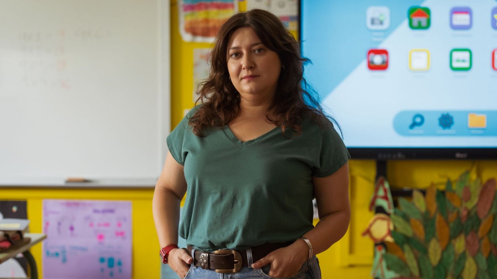 Mujer con camiseta verde y vaqueros, de pie frente a una pared amarilla, con una pizarra y una pantalla digital interactiva.