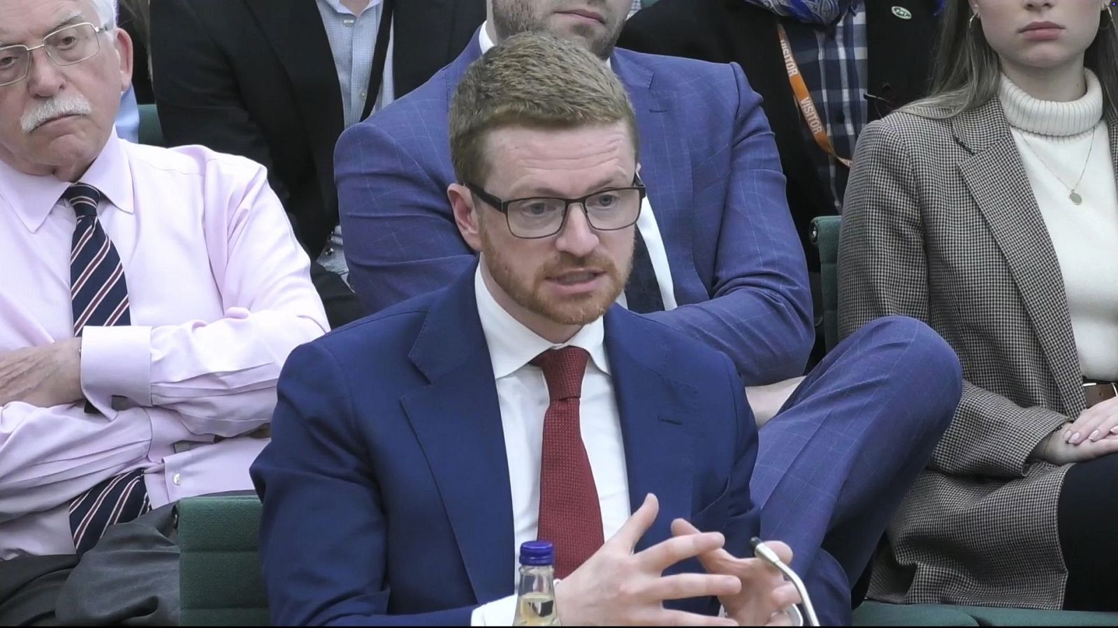 Morgan McSweeney, exjefe de gabinete de Downing Street, comparece ante la Comisión de Asuntos Exteriores. Se le ve hablando, con traje azul, camisa blanca y corbata roja, gafas y barba pelirroja. A su lado, un hombre mayor y una mujer joven escuchan.